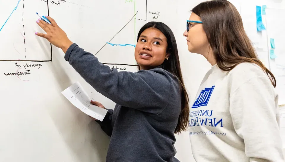 A U N E student points to a graph on a white board